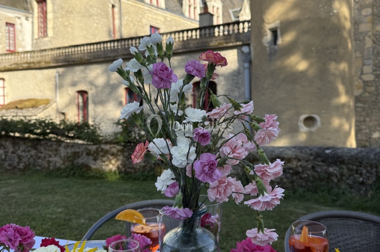 Vase de fleurs roses et blanches sur une table en extérieur, devant un bâtiment en pierre avec des fenêtres rouges.