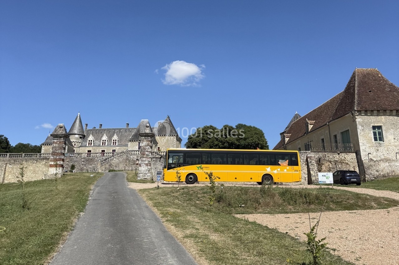 Autocar jaune stationné devant un château ancien entouré de murs en pierre, sous un ciel bleu dégagé.