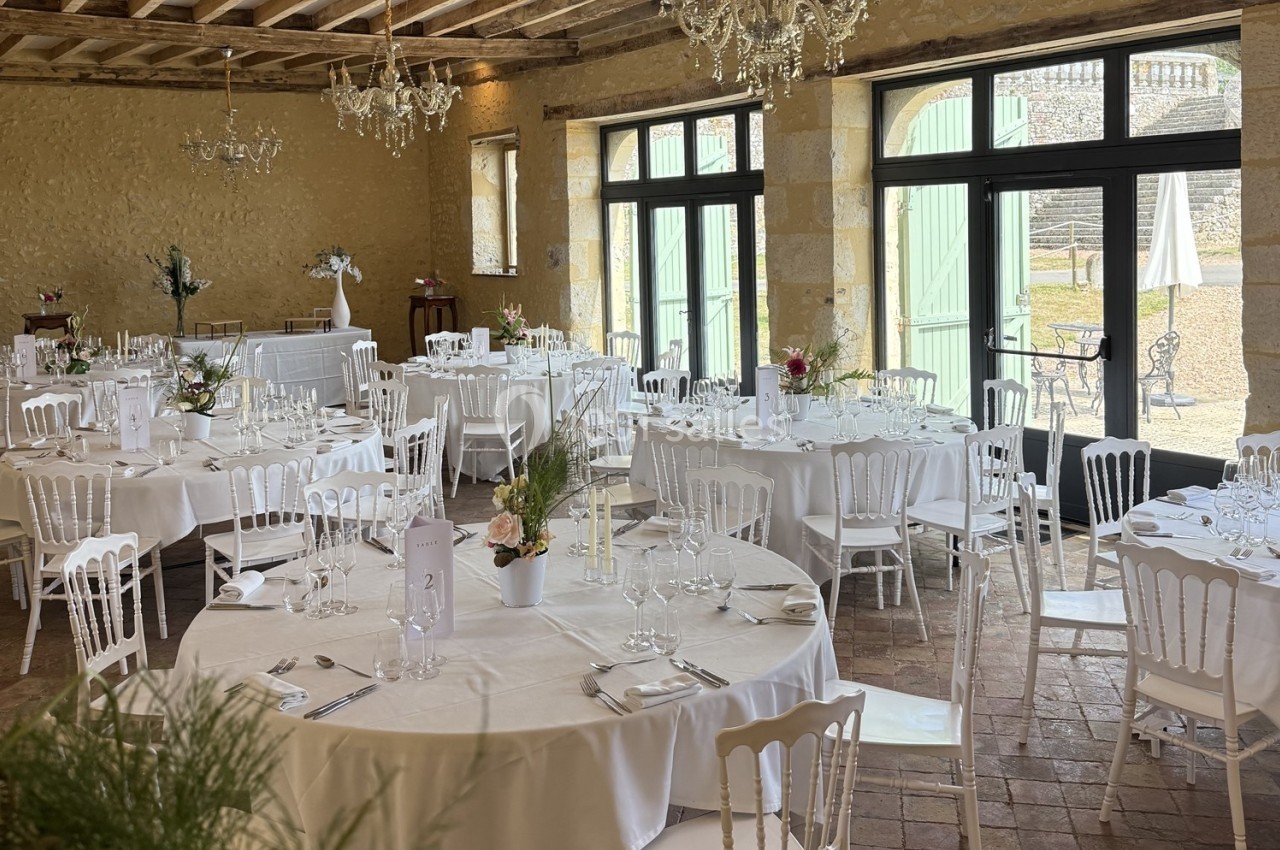 Salle de réception avec tables rondes dressées, nappes blanches, chaises en bois et lumière naturelle entrant par de grandes…
