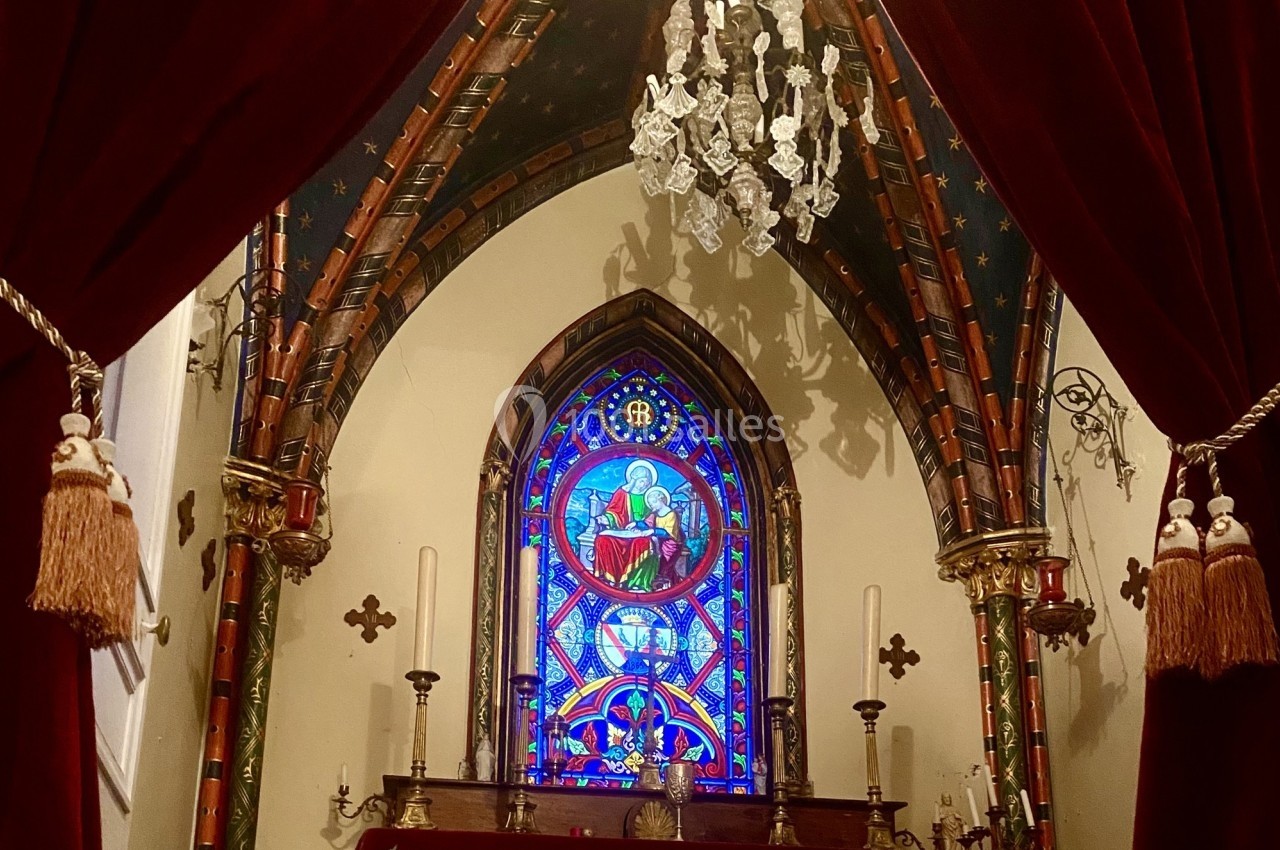 Vitrail coloré représentant une scène religieuse dans une chapelle ornée de rideaux rouges et d'un lustre.