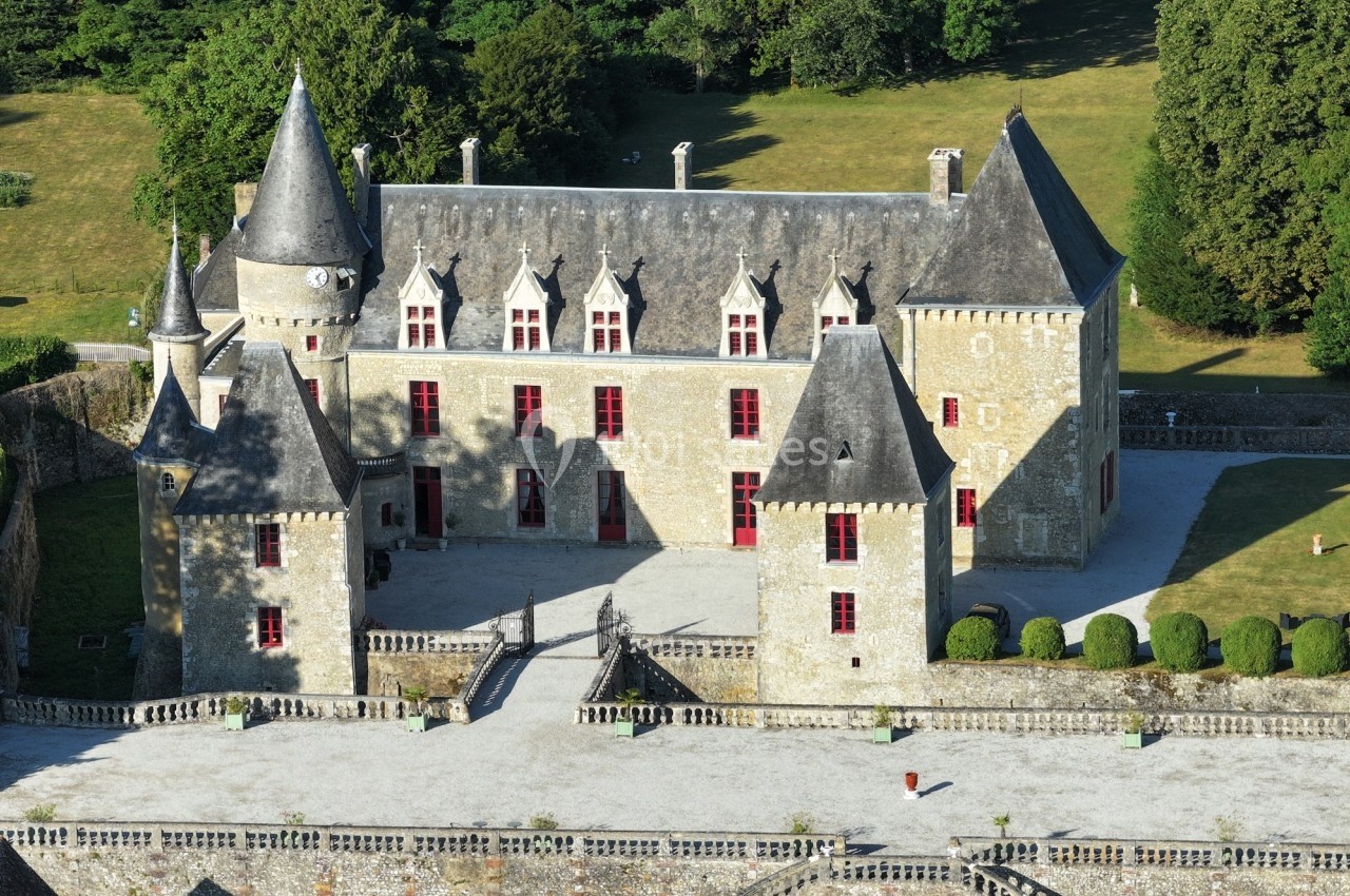 Château en pierre avec quatre tours et des fenêtres rouges, entouré de verdure et d'une cour pavée.