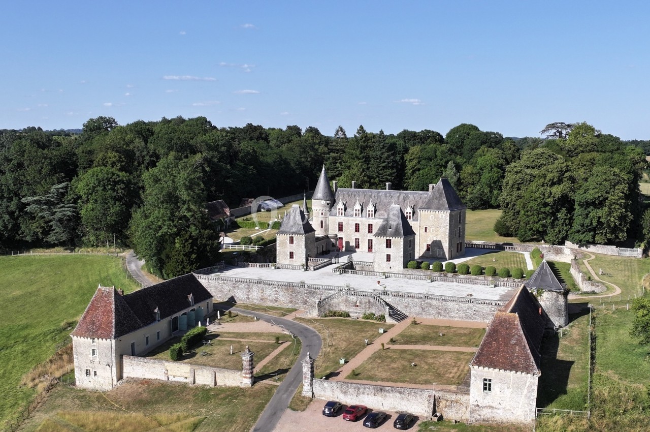 Vue aérienne d'un château entouré de verdure, avec des dépendances, une cour centrale et des voitures stationnées.
