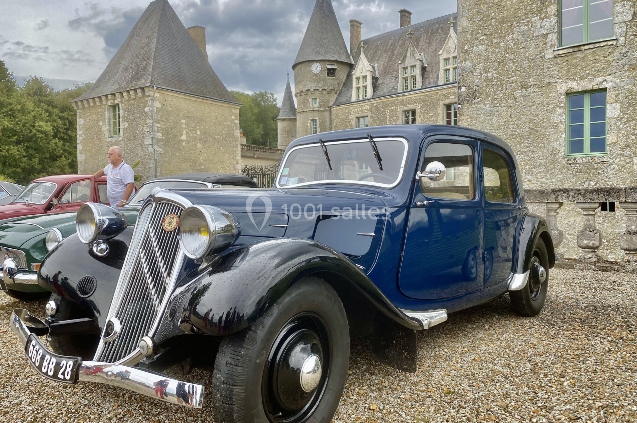 Voiture ancienne bleue et noire stationnée devant un château en pierre, avec un ciel nuageux en arrière-plan.