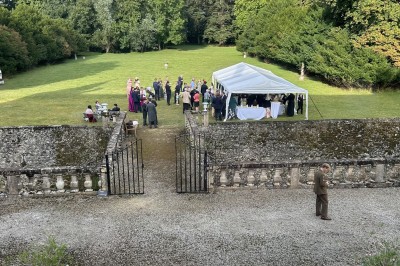 Château éclairé de nuit avec des lumières colorées, entouré de buissons et de tentes blanches sous un ciel nuageux.