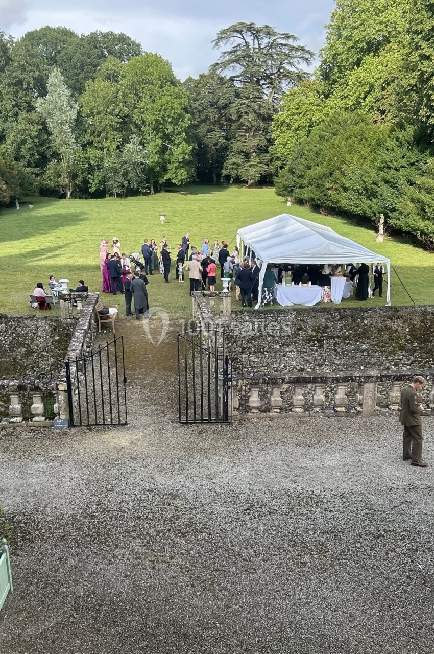 Groupe de personnes rassemblées près d'une tente blanche dans un jardin entouré d'arbres et d'une allée gravillonnée.