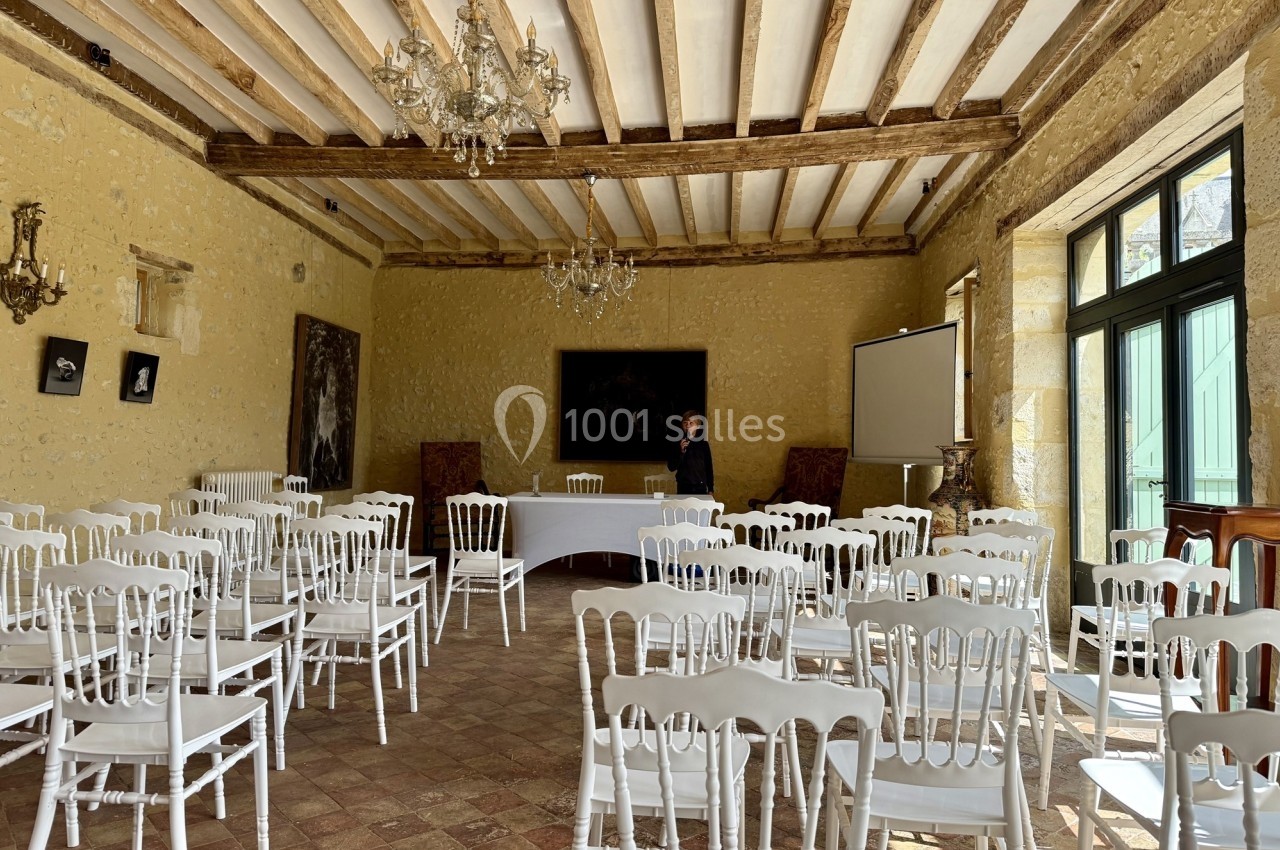 Salle lumineuse avec des chaises blanches alignées, un écran de projection et un homme debout près d'une table.