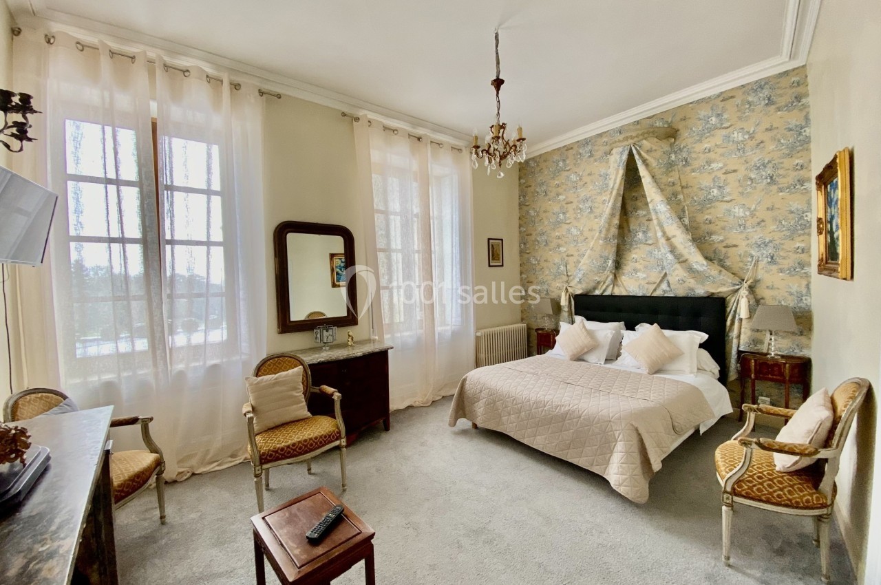 Chambre lumineuse avec lit double, mobilier ancien, murs clairs, rideaux transparents et vue sur l'extérieur.