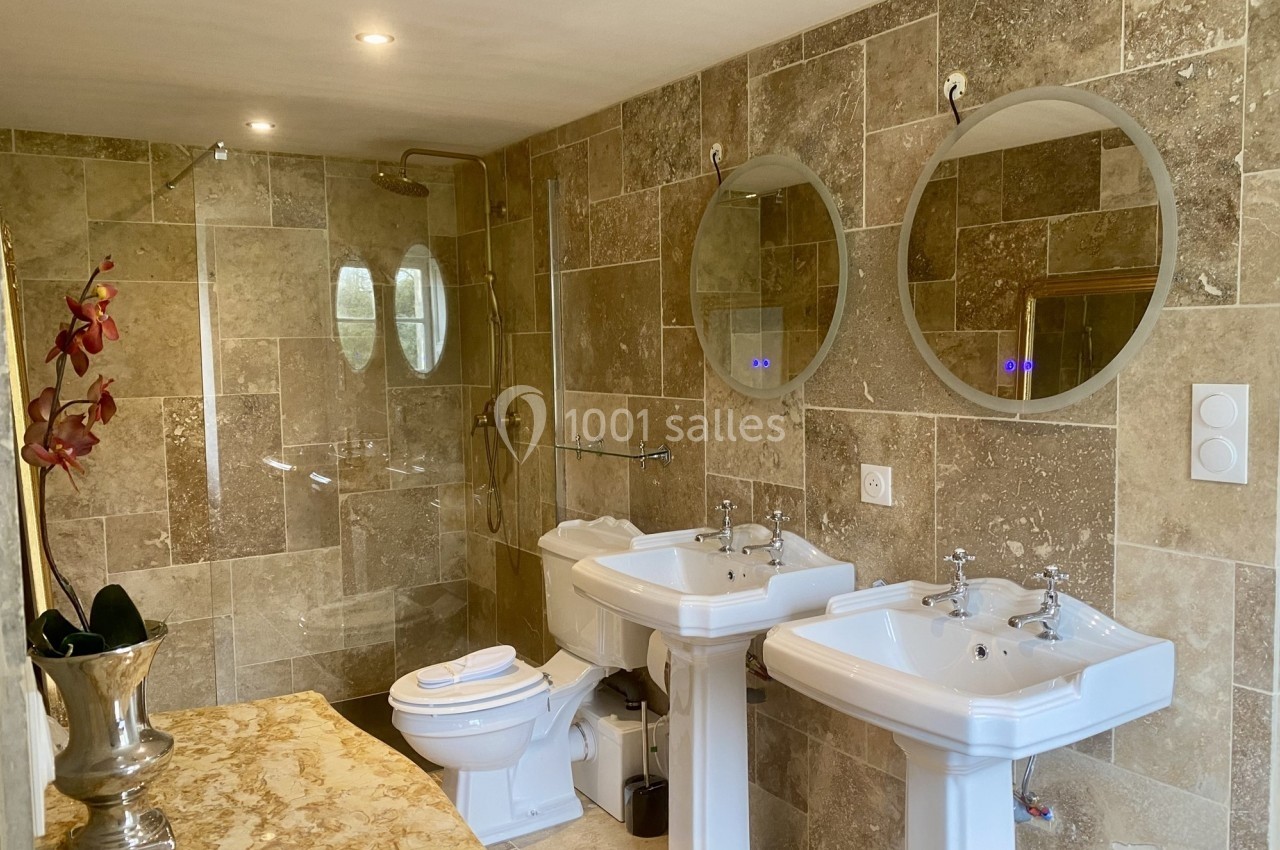 Salle de bain avec murs en pierre beige, deux lavabos sur pied, une douche, un WC et des miroirs ronds.