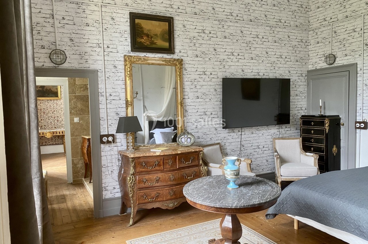 Chambre élégante avec murs en briques blanches, mobilier classique, miroir doré et télévision murale.