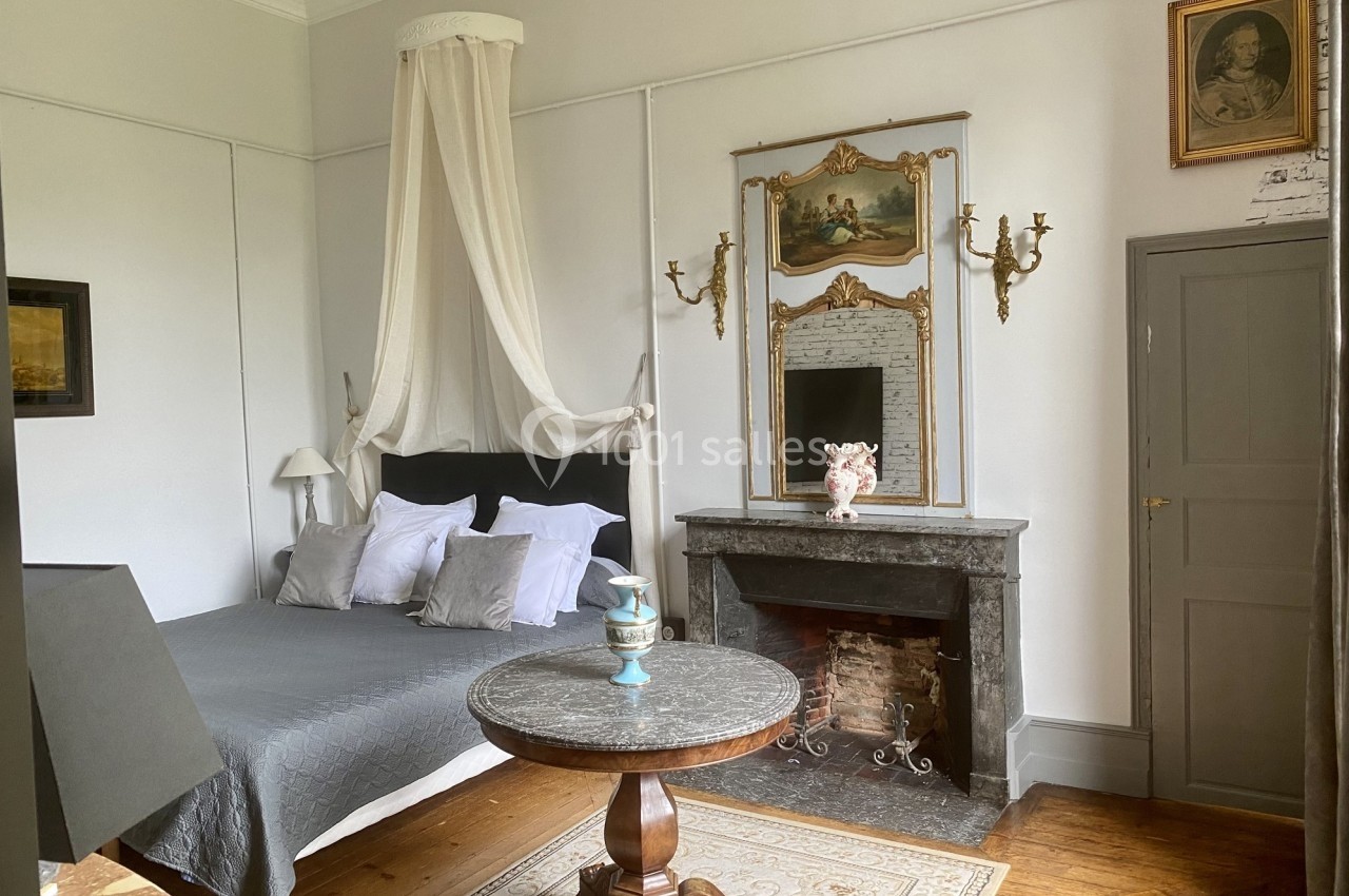 Chambre élégante avec lit à baldaquin, cheminée en marbre, miroir doré et table ronde sur un tapis.