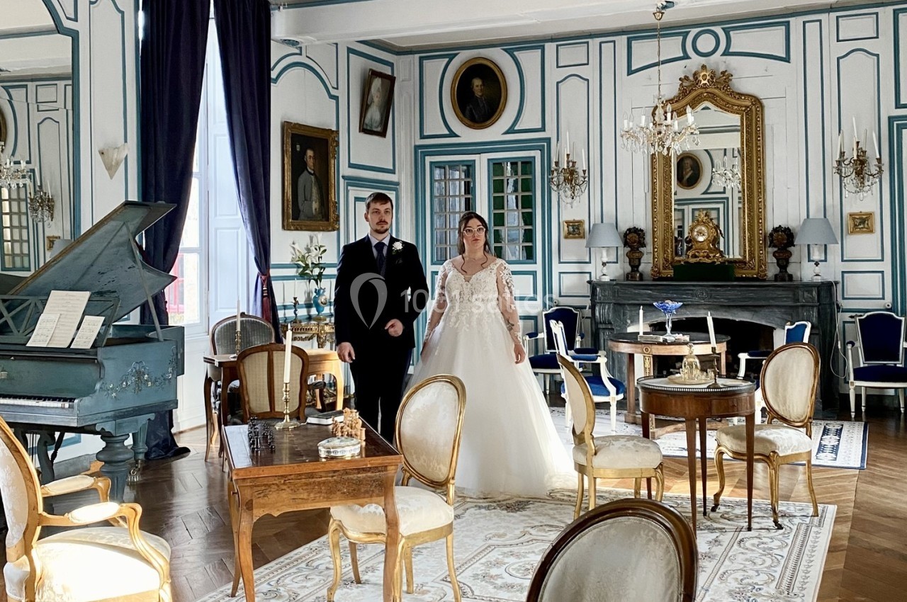Un couple en tenue de mariage se tient dans un salon élégant avec piano, miroirs et mobilier ancien.