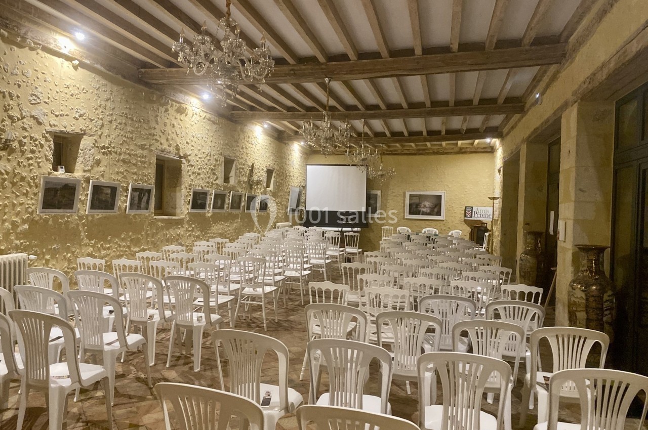 Salle en pierre avec des rangées de chaises blanches disposées face à un écran de projection.