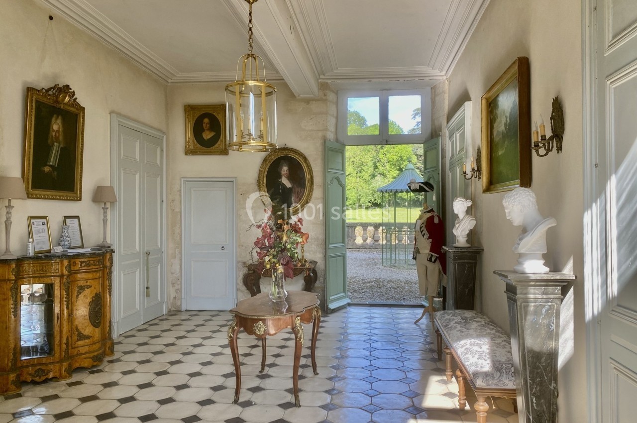 Intérieur d'une entrée lumineuse avec sol en damier, meubles anciens, portraits et vue sur un jardin.