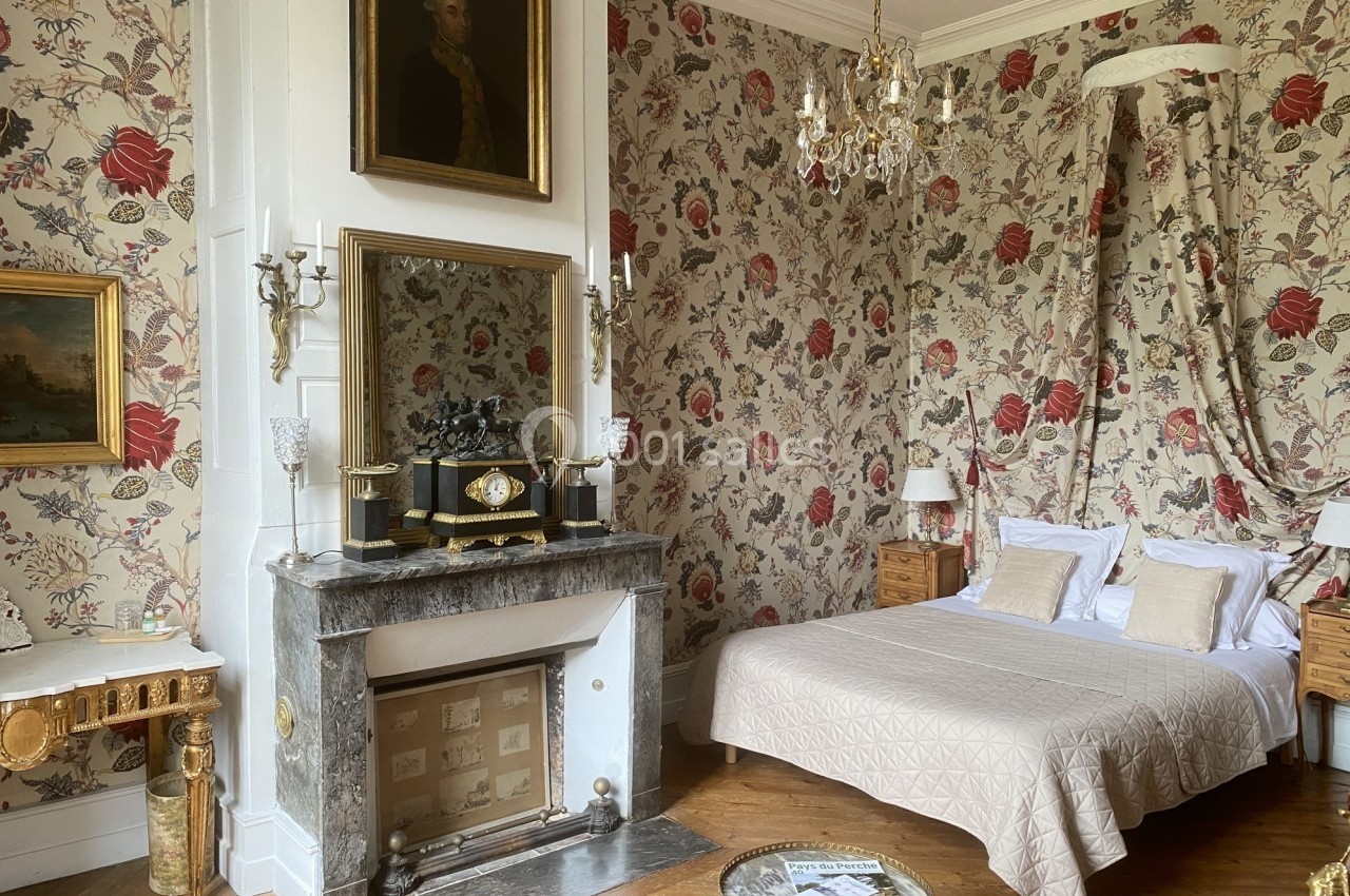 Chambre élégante avec lit double, cheminée en marbre, lustre et murs décorés de papier peint fleuri.
