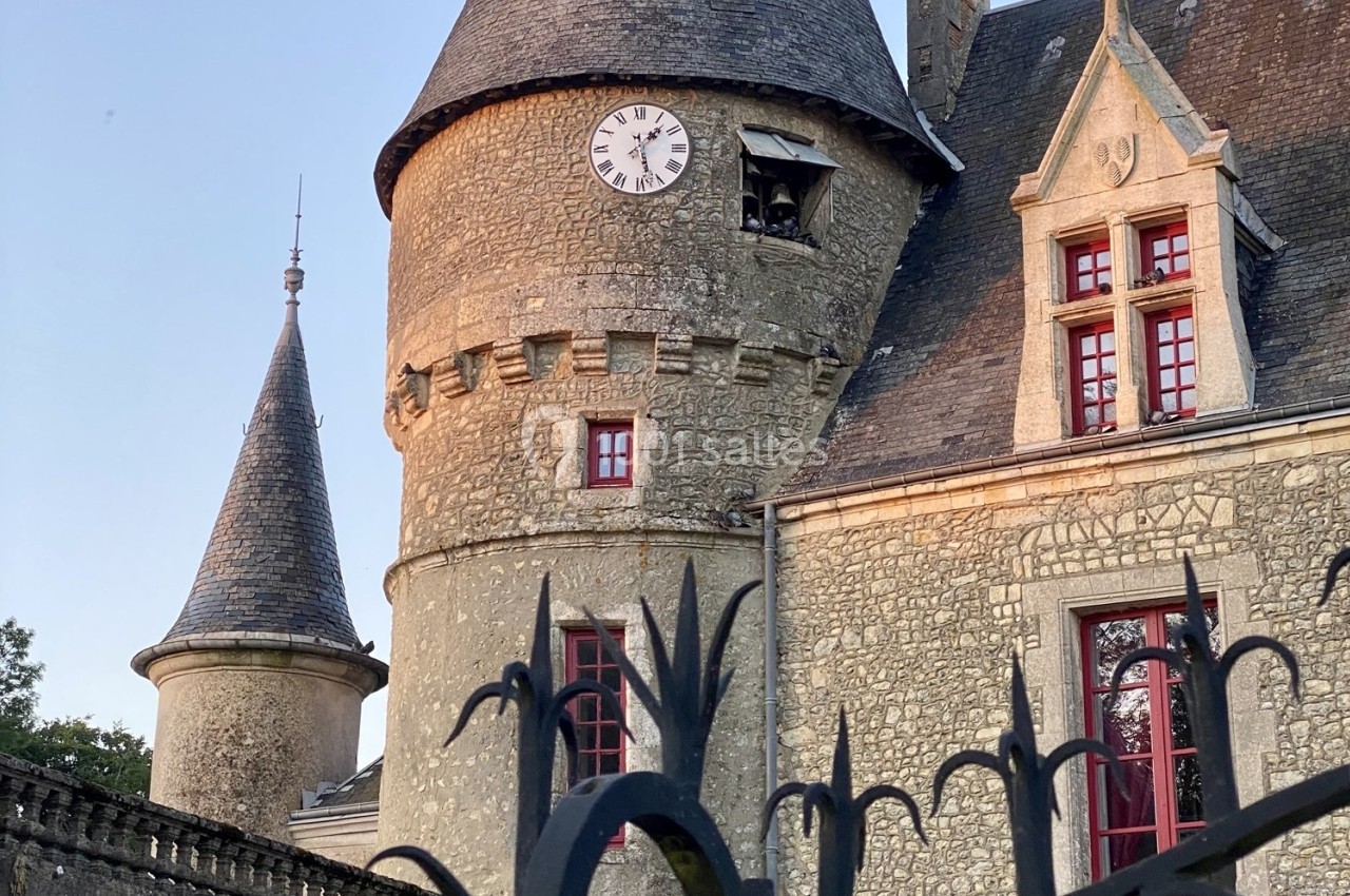 Tour en pierre d'un château avec horloge, fenêtres rouges et toit en ardoise, vue derrière une grille noire.