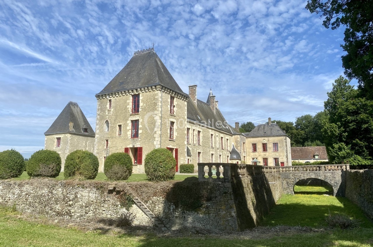 Château en pierre avec toits en ardoise, entouré de buissons taillés, d'un pont et d'un ciel partiellement nuageux.
