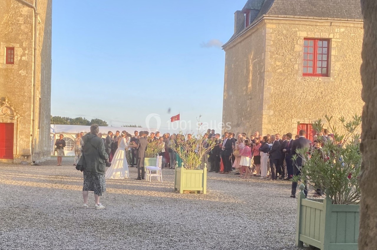 Groupe de personnes rassemblées dans une cour ensoleillée entre deux bâtiments en pierre avec des fenêtres rouges.