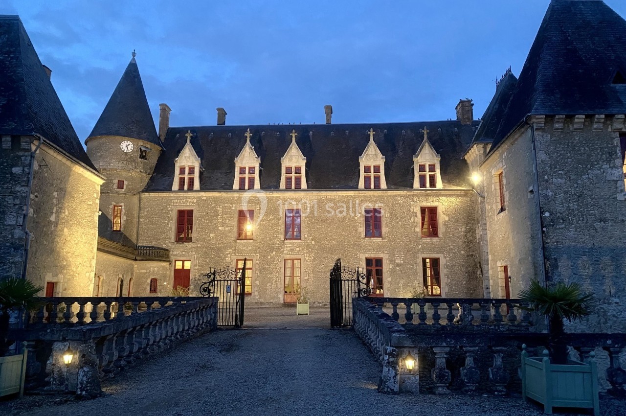 Façade d'un château en pierre éclairé à la tombée de la nuit, avec une cour et des balustrades au premier plan.
