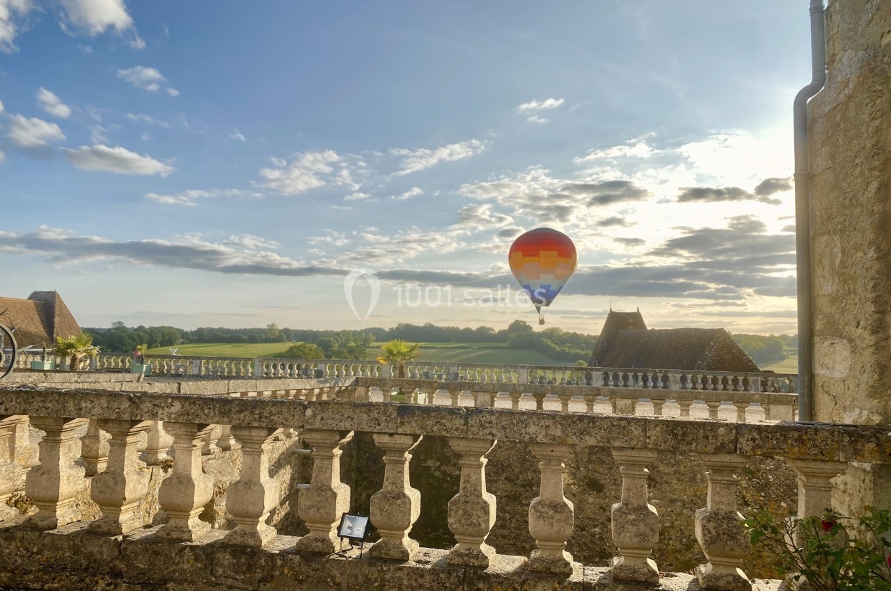 Montgolfière colorée flottant dans un ciel partiellement nuageux, vue depuis une terrasse en pierre avec balustrade.
