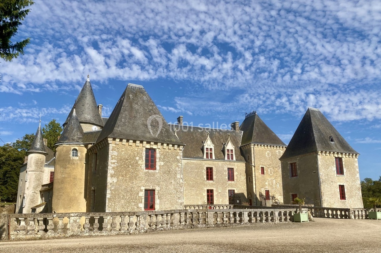 Façade d'un château en pierre avec plusieurs tours et fenêtres rouges, sous un ciel partiellement nuageux.