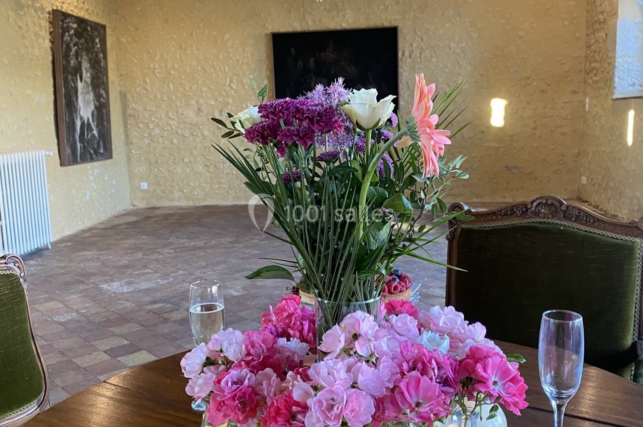 Vase de fleurs colorées sur une table en bois, entouré de verres, dans une pièce aux murs en pierre.
