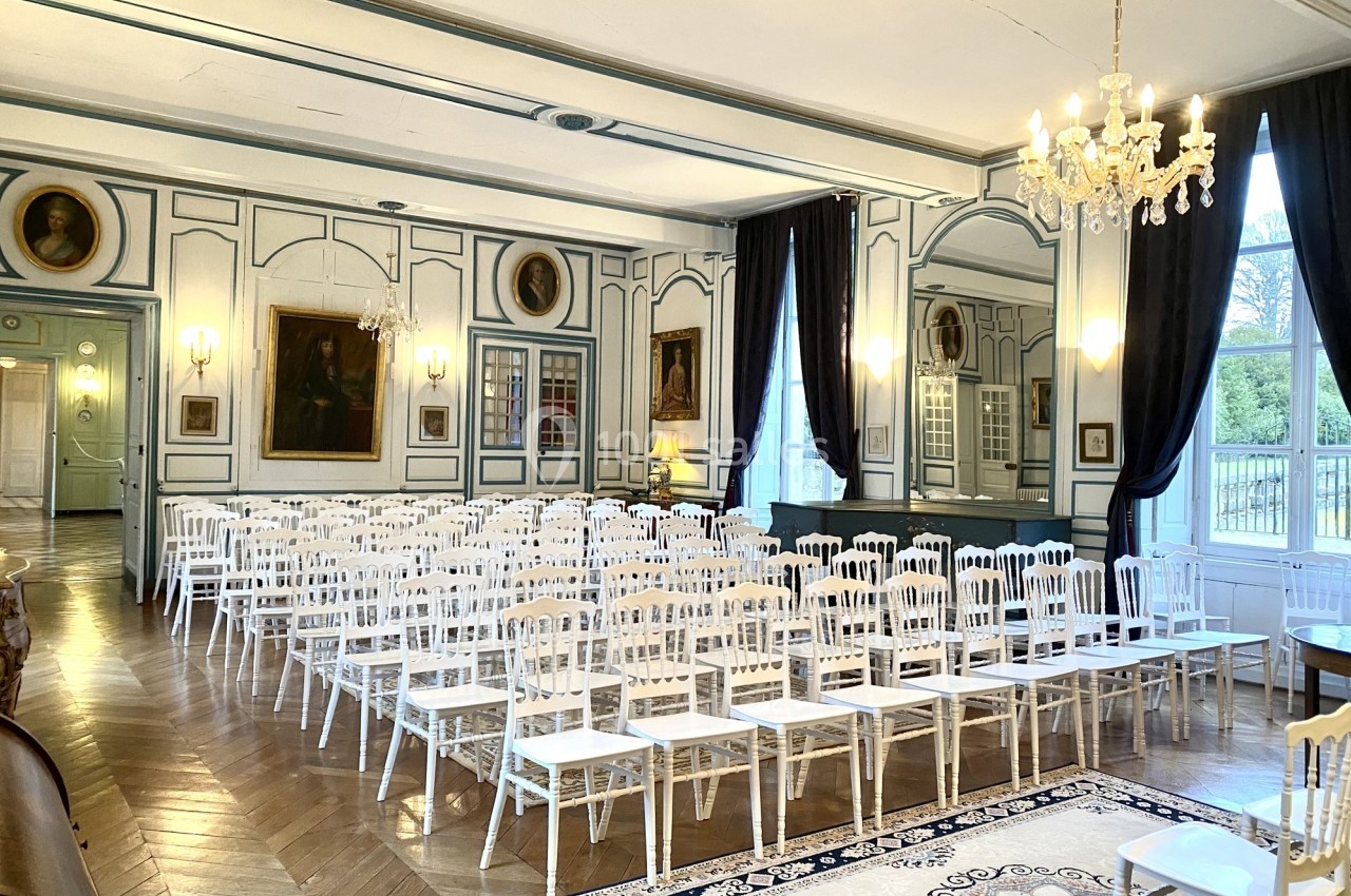 Salle élégante avec parquet, chaises blanches alignées, lustres et peintures encadrées sur des murs ornés de moulures.