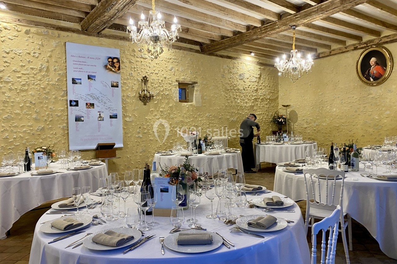 Salle de réception avec tables rondes dressées, décor rustique, chandeliers et une personne préparant le service.
