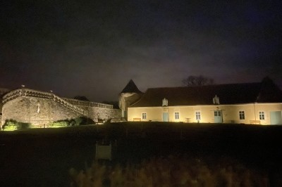 Château éclairé de nuit avec des lumières colorées, entouré de buissons et de tentes blanches sous un ciel nuageux.