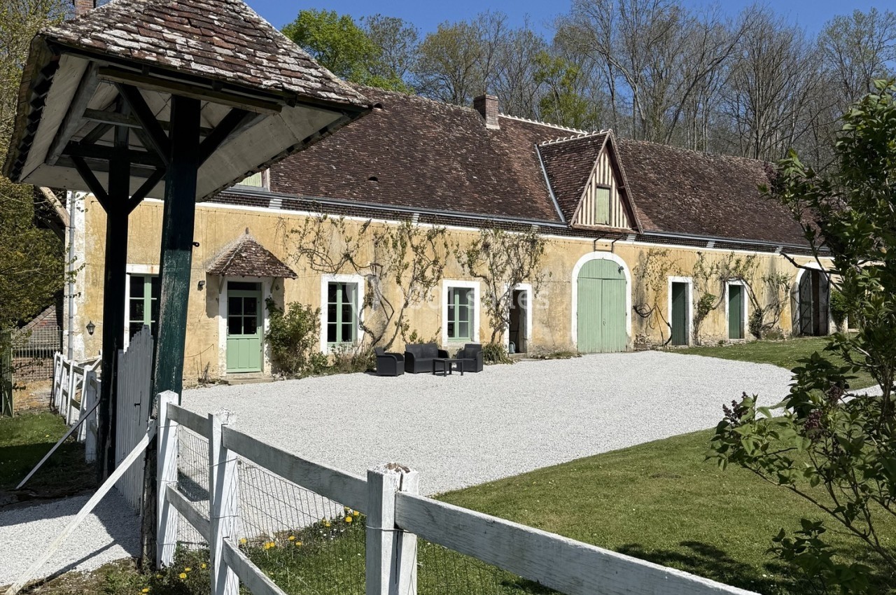 Façade d'une maison ancienne en pierre avec toit en tuiles, entourée de verdure et d'une cour gravillonnée.