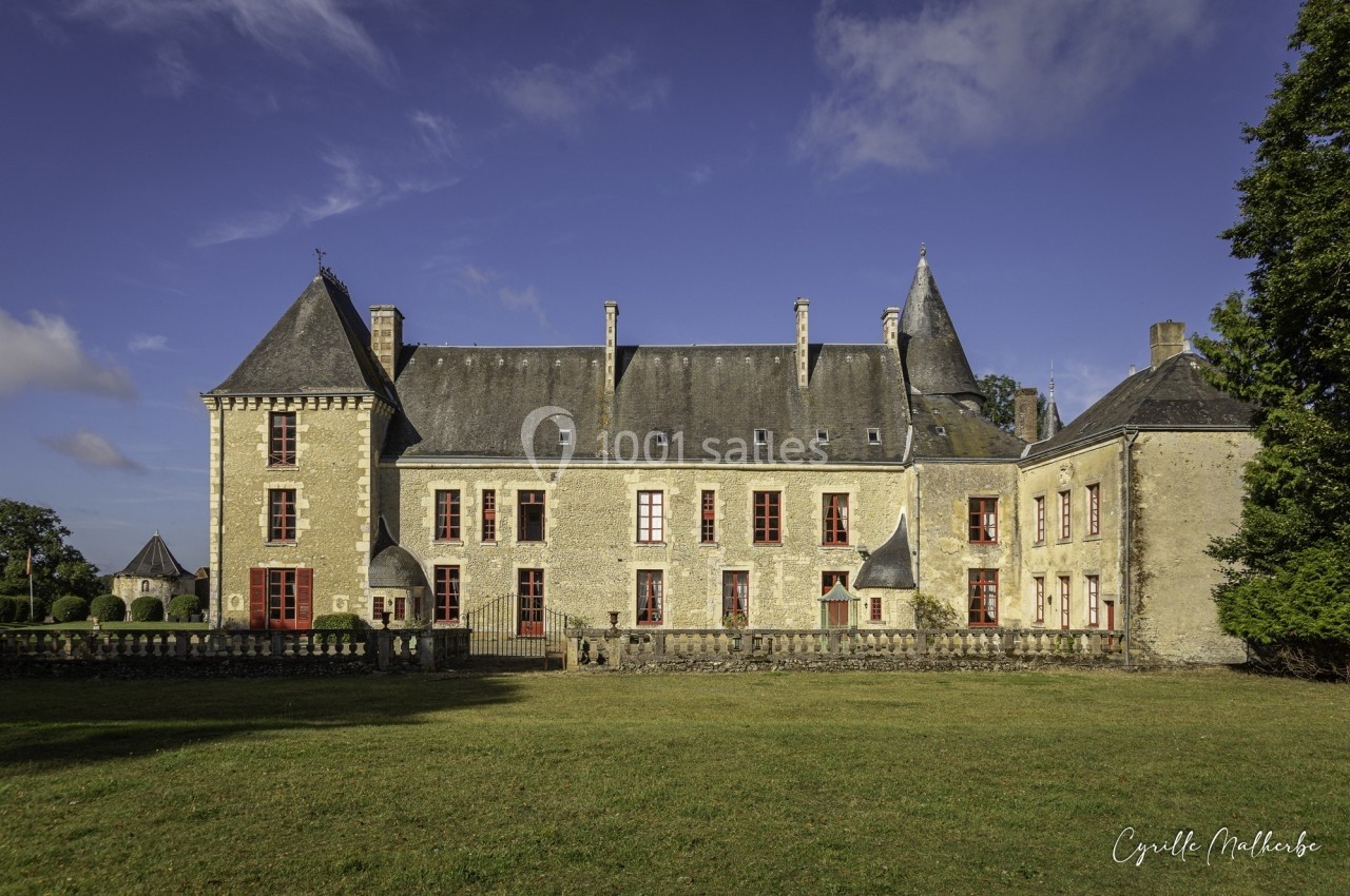 Façade d'un château en pierre avec plusieurs tours et fenêtres rouges, entouré d'une pelouse et d'arbres.