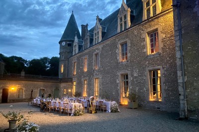Château éclairé de nuit avec des lumières colorées, entouré de buissons et de tentes blanches sous un ciel nuageux.