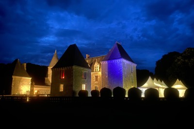 Château éclairé de nuit avec des lumières colorées, entouré de buissons et de tentes blanches sous un ciel nuageux.