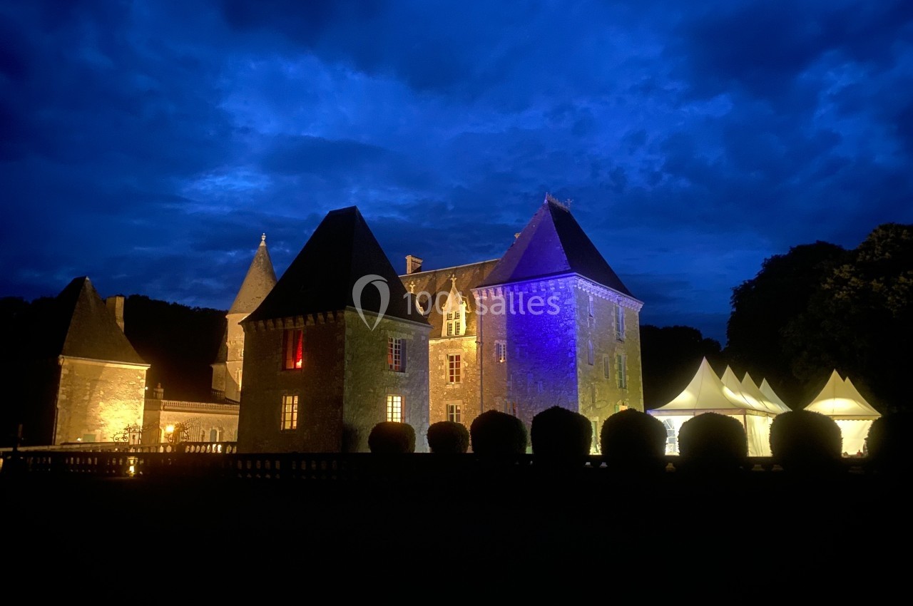 Château éclairé de nuit avec des lumières colorées, entouré de buissons et de tentes blanches sous un ciel nuageux.