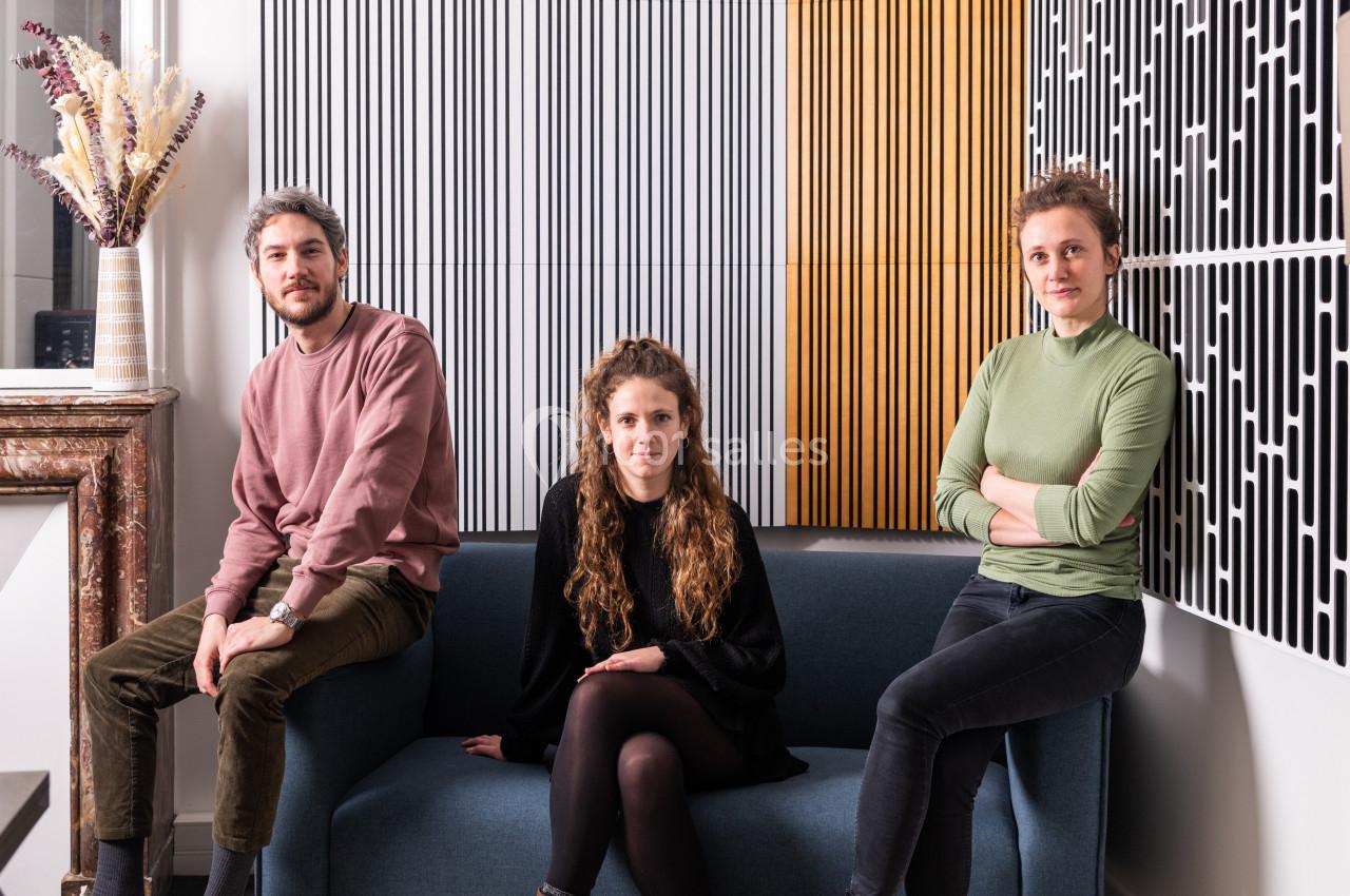 Trois personnes assises et debout dans un intérieur moderne avec des panneaux muraux décoratifs en arrière-plan.