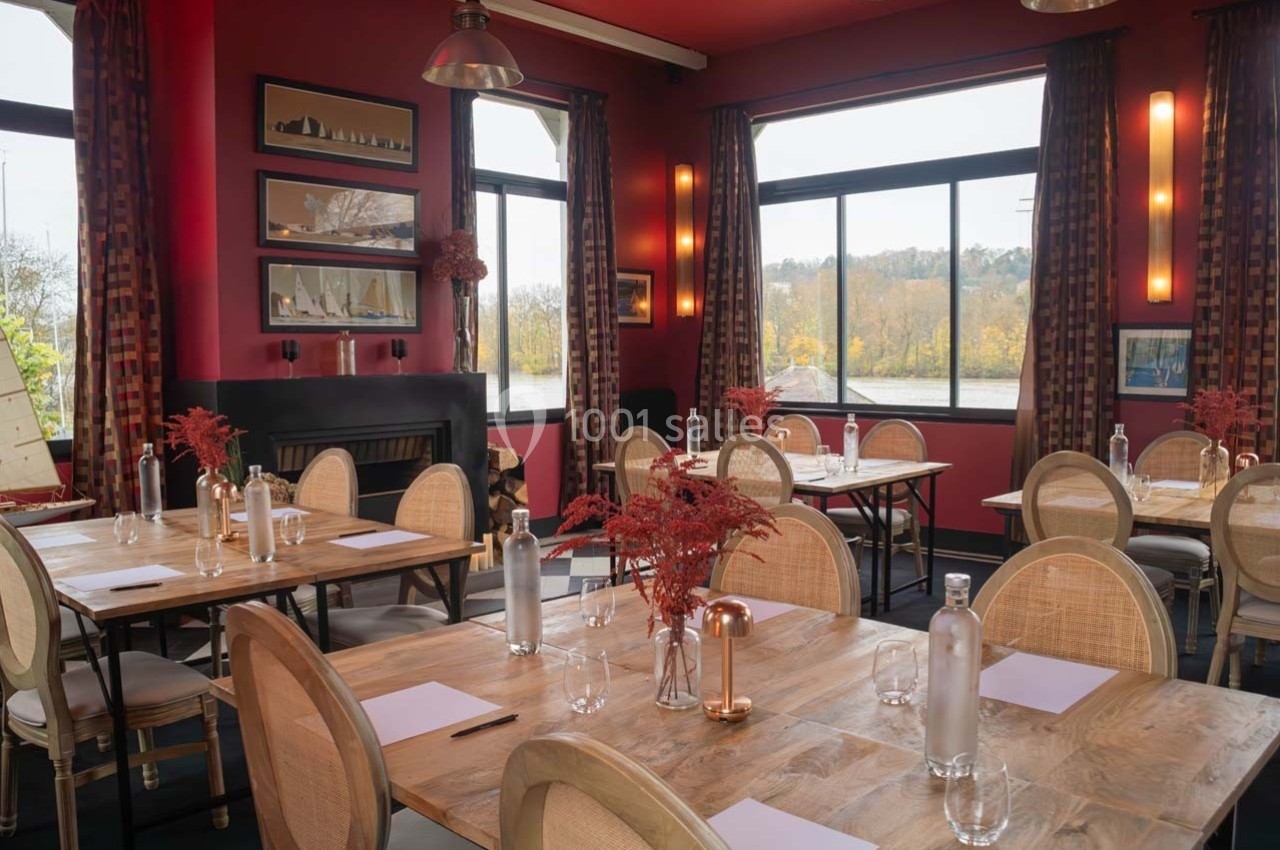 Salle de restaurant lumineuse avec tables en bois, chaises en rotin, décor rouge et grandes fenêtres donnant sur un paysage.