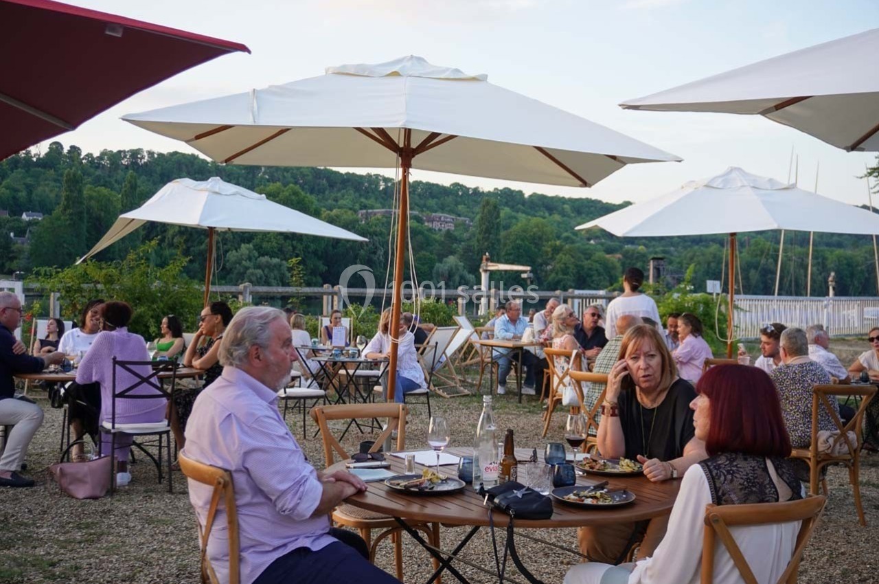 Personnes dînant en terrasse sous des parasols, avec vue sur un paysage verdoyant en arrière-plan.
