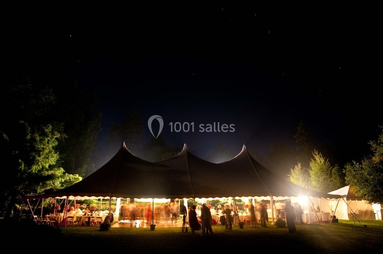 Grande tente éclairée la nuit, entourée de personnes et d'arbres, dans un cadre extérieur sombre.