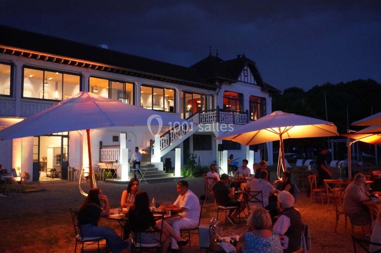 Personnes dînant en terrasse sous des parasols éclairés, devant un bâtiment illuminé au crépuscule.