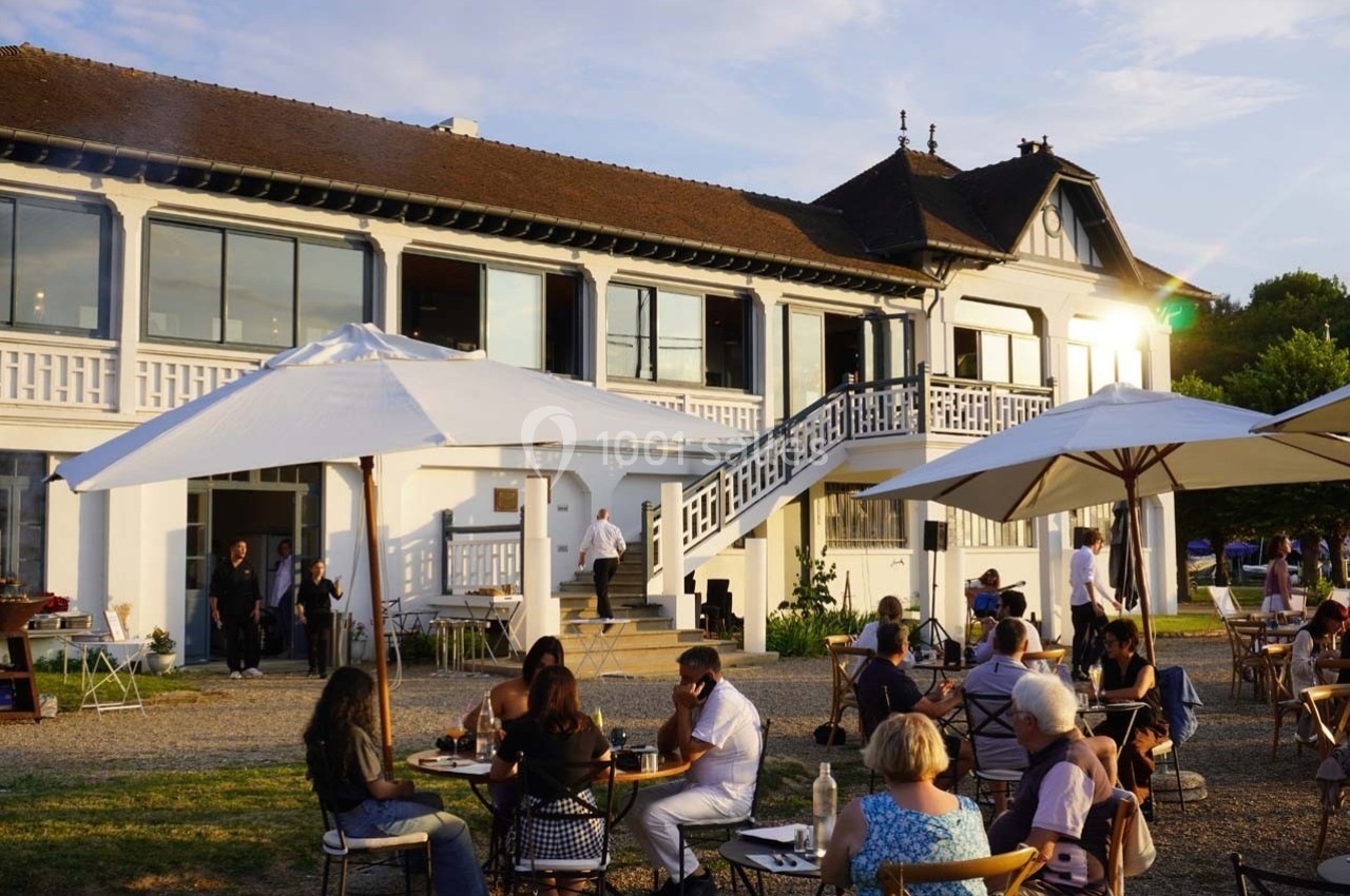 Personnes attablées en terrasse devant un grand bâtiment blanc avec baies vitrées, par une fin de journée ensoleillée.
