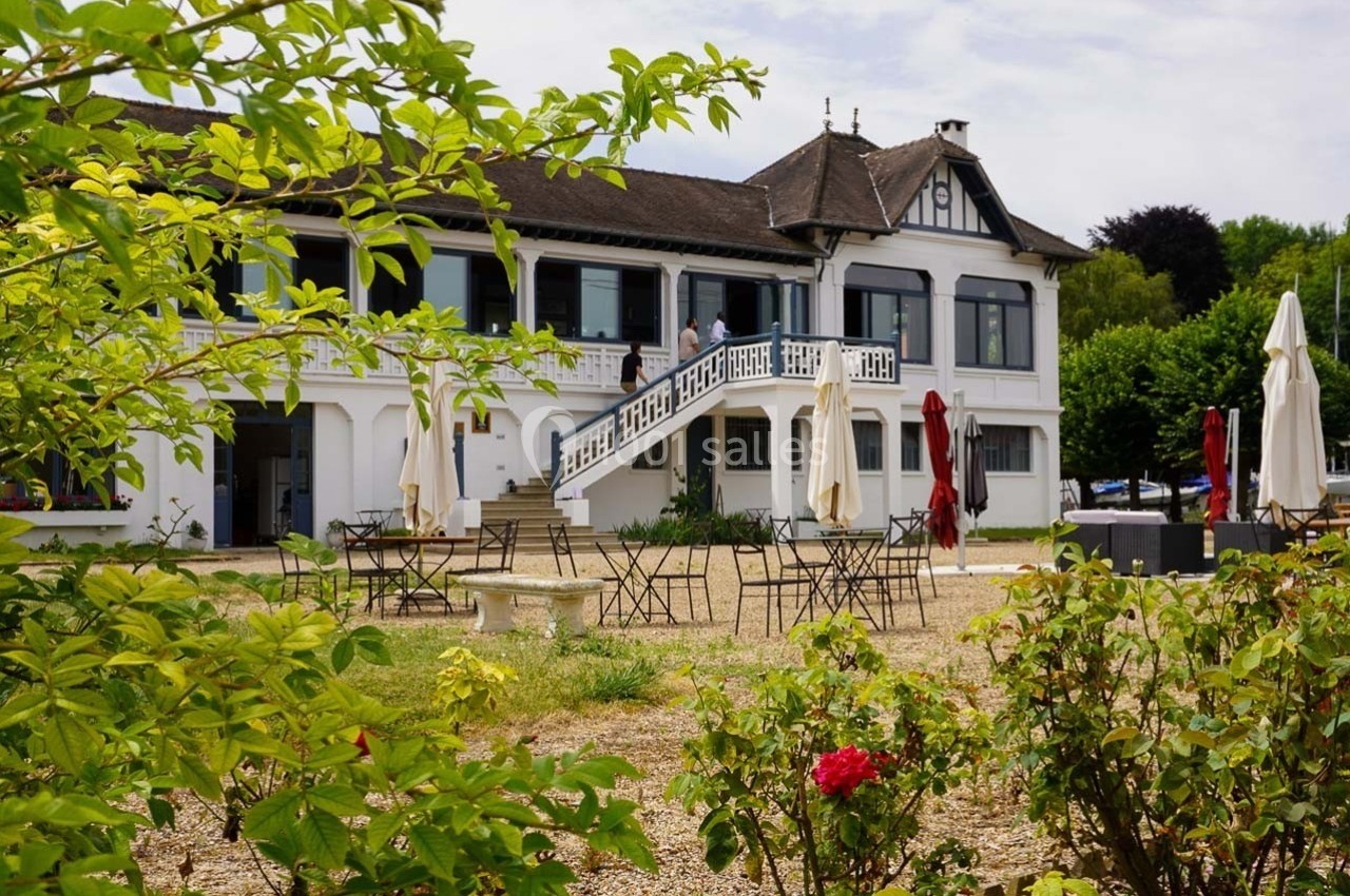 Bâtiment blanc à deux étages avec terrasse, entouré de tables, chaises et végétation sous un ciel clair.