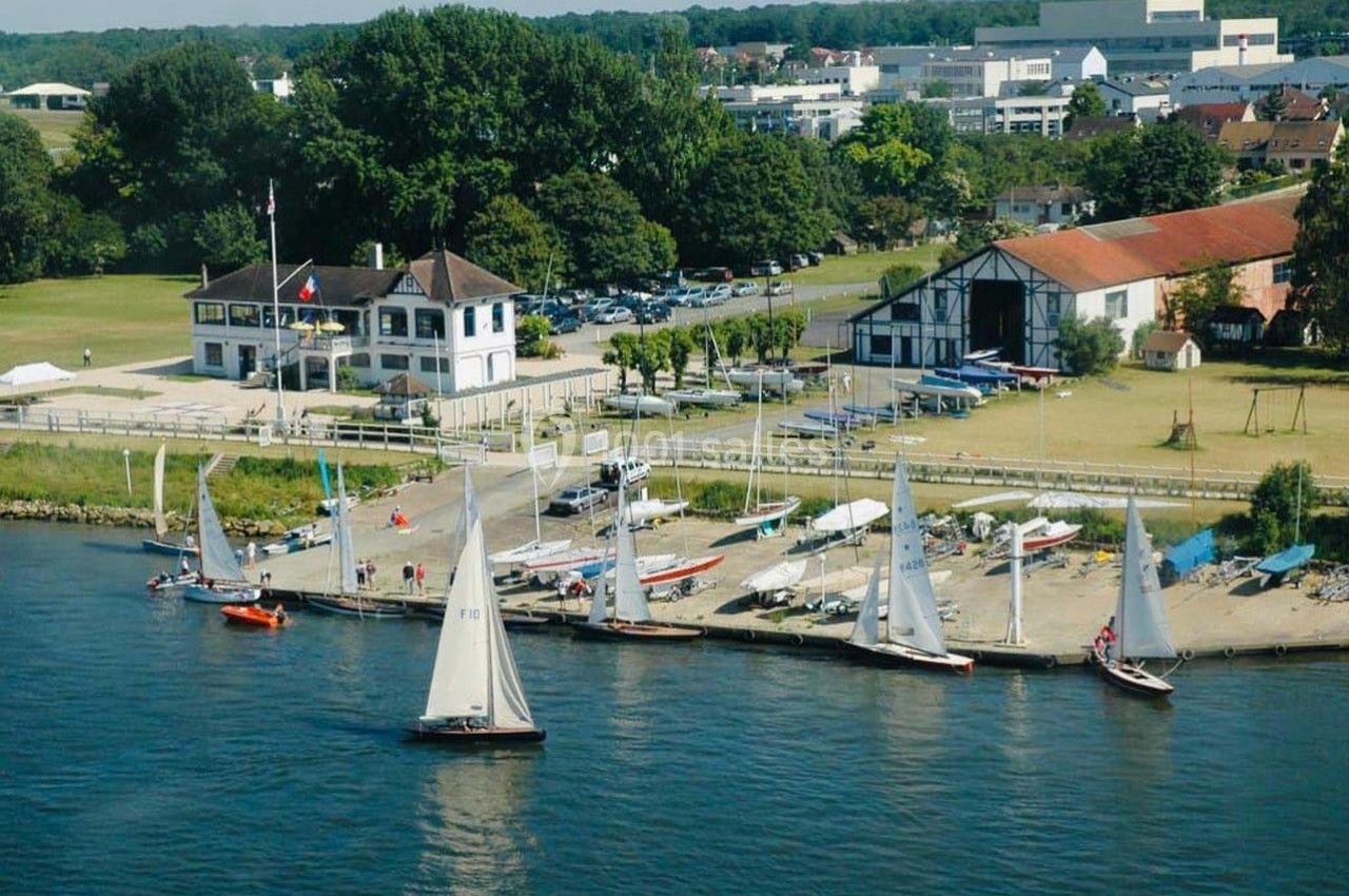 Vue aérienne d'un port de plaisance avec des voiliers amarrés, des bâtiments et un espace vert en arrière-plan.