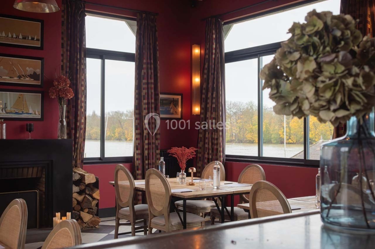 Salle de restaurant chaleureuse avec murs rouges, grandes fenêtres donnant sur un paysage automnal et décoration soignée.