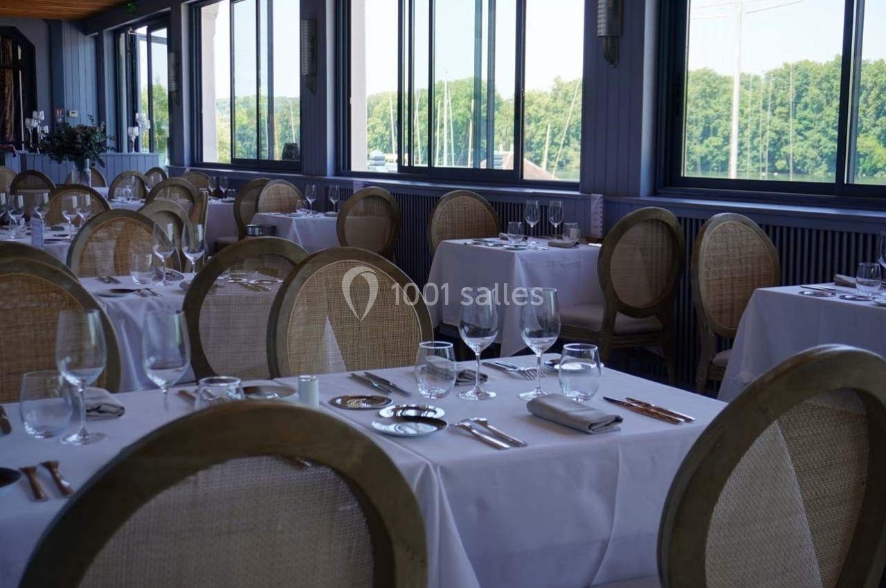 Salle de restaurant lumineuse avec tables dressées, nappes blanches et grandes fenêtres donnant sur un paysage verdoyant.