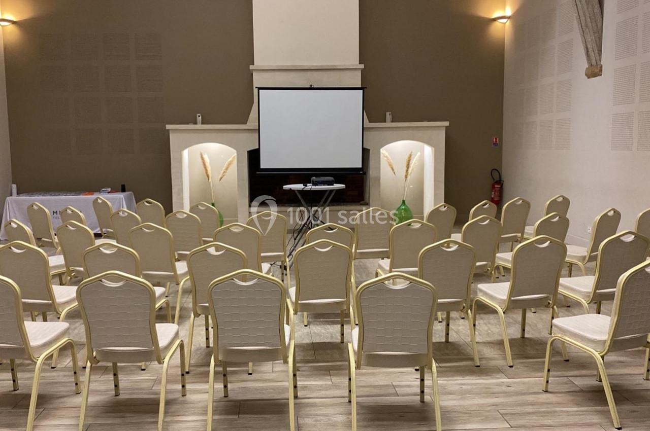 Salle de conférence avec des rangées de chaises beige alignées face à un écran de projection et un pupitre.