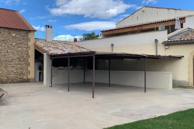 Abri extérieur avec toit en tuiles et structure métallique, situé sur une terrasse en béton près d'un bâtiment en pierre.