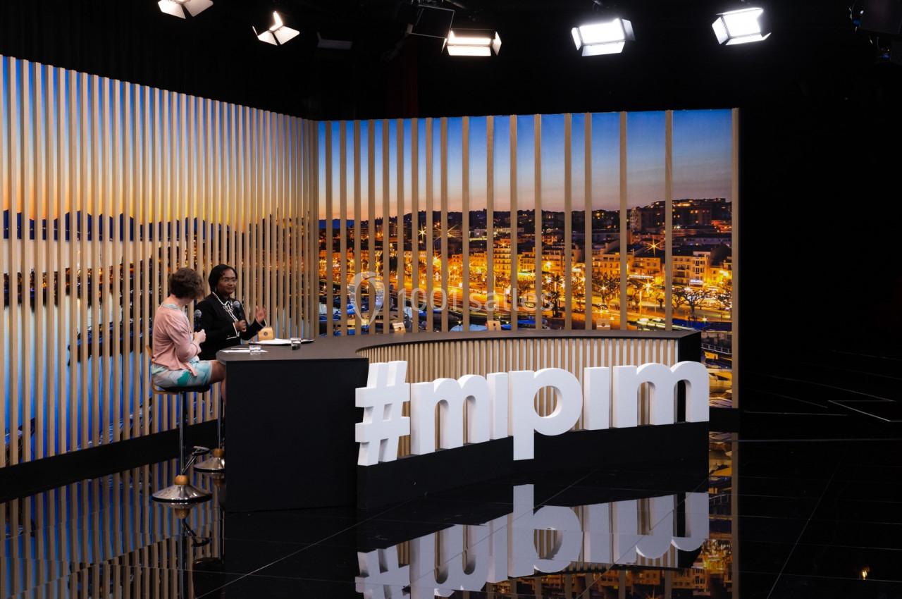 Deux personnes discutent sur un plateau télévisé avec vue nocturne sur une ville en arrière-plan.