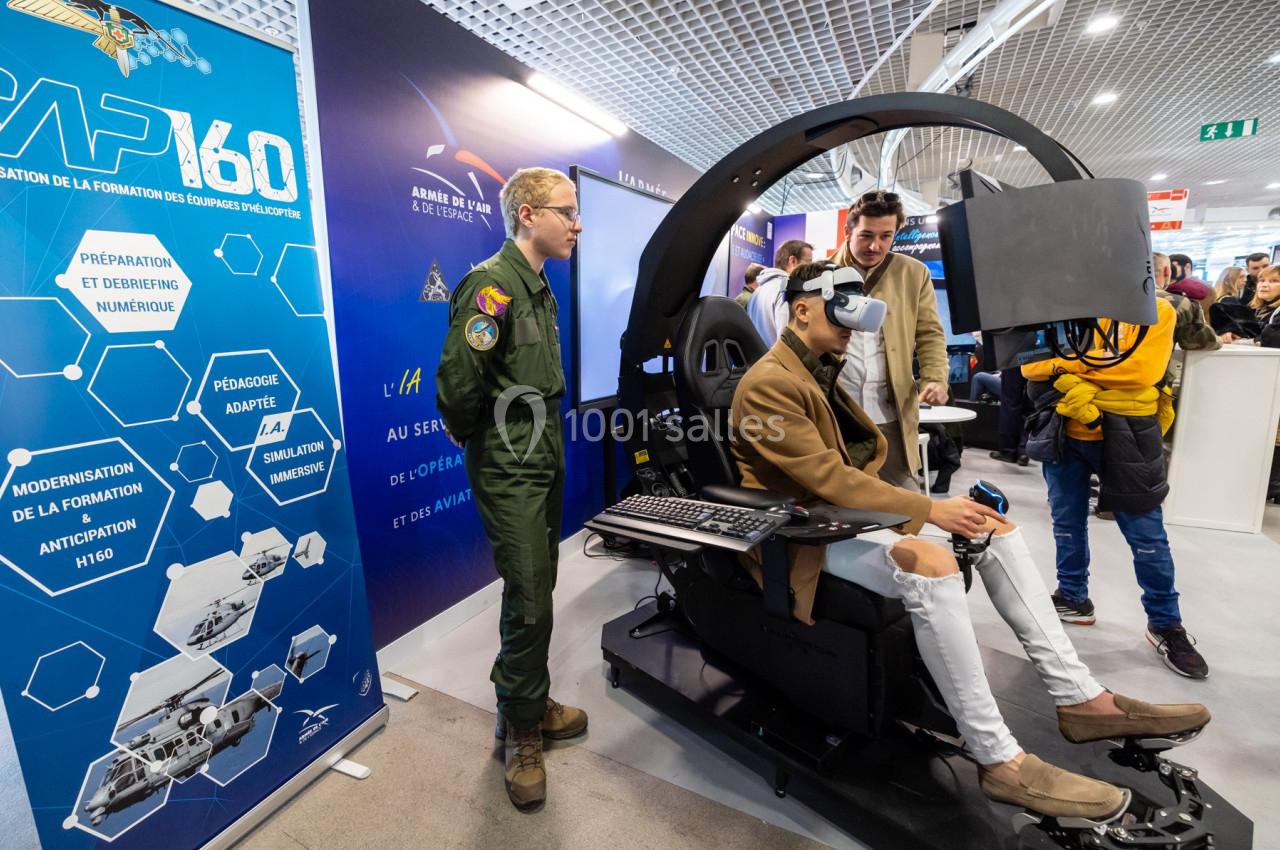 Un homme utilise un simulateur de vol avec un casque de réalité virtuelle, entouré de spectateurs et d'un instructeur en…