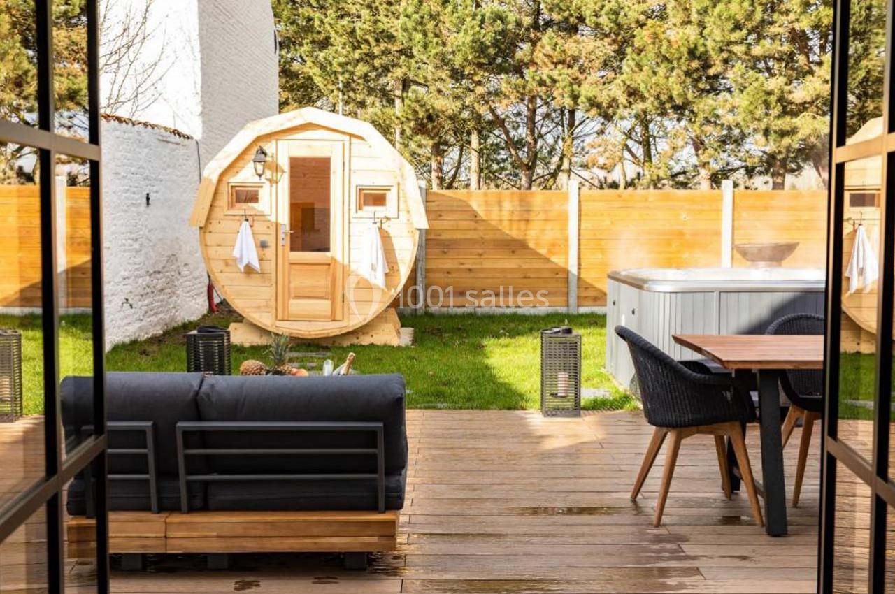 Terrasse en bois avec salon de jardin, sauna en forme de tonneau, jacuzzi et clôture en bois, entourée de verdure.