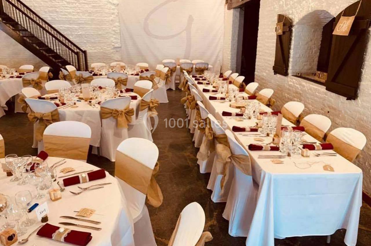 Salle de réception décorée avec des tables dressées, nappes blanches, serviettes rouges et chaises ornées de nœuds beiges.