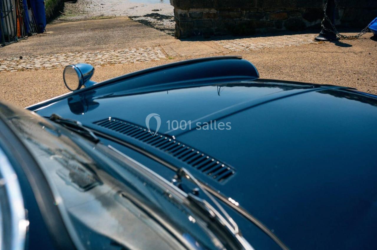 Capot d'une voiture classique bleu foncé avec une prise d'air, stationnée près d'un chemin pavé et d'un plan d'eau.